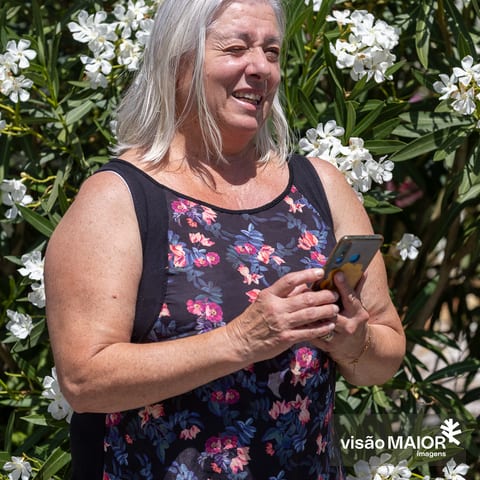 Mulher em pé, cabelo branco a semi cerrar os olhos, de telemóvel na mão