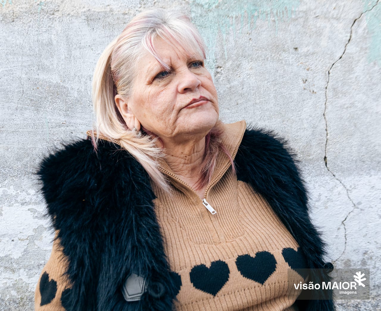 Mulher com ar sério, cabelo e olhos claros, com um colete de pelo preto e camisola clara com corações