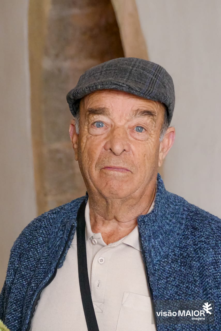 Homem com ar sério, olhos claros e boné, casaco de malha azul e polo branca