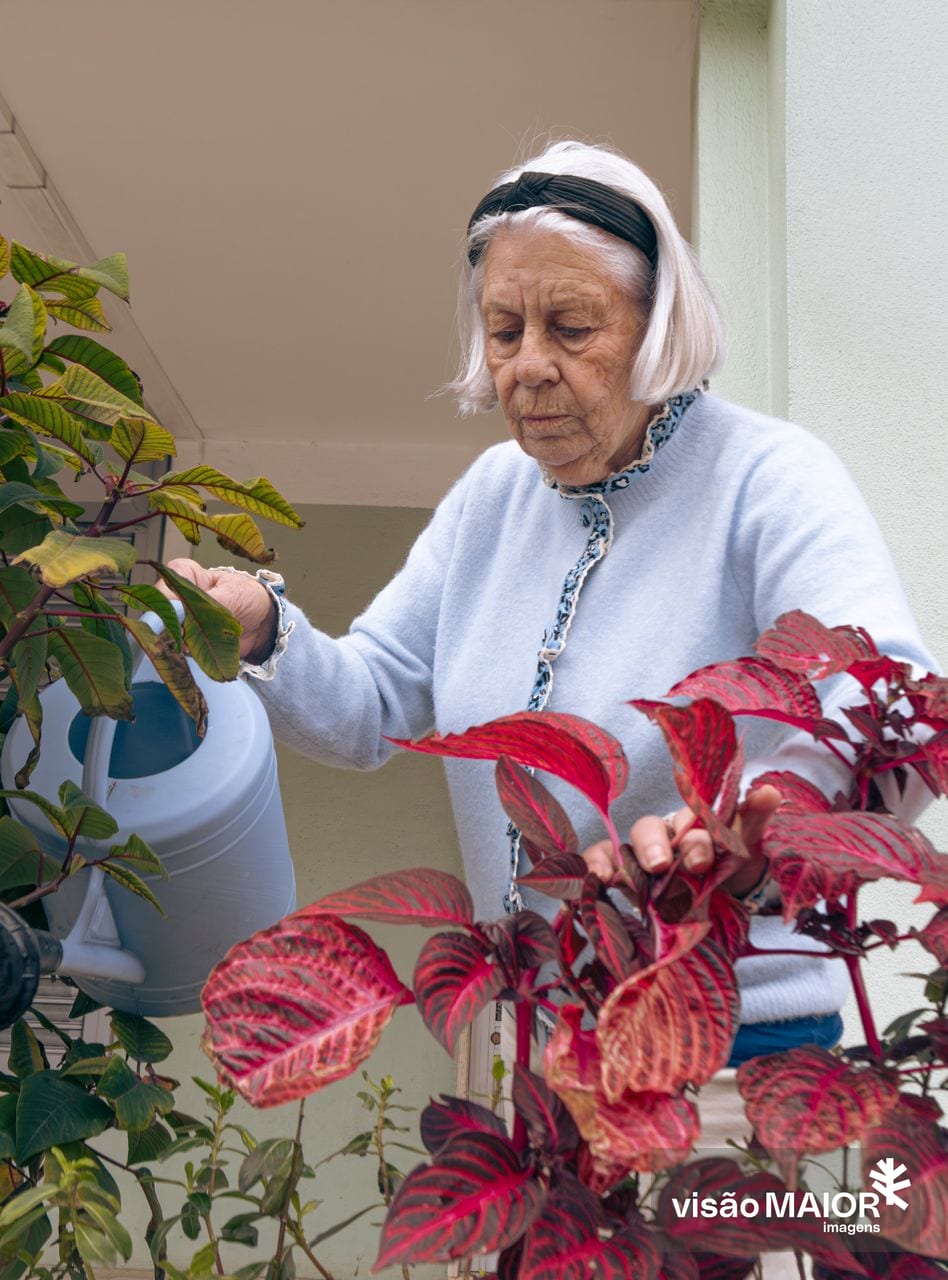 Senhora de cabelo branco, fita na cabeça e casaco claro com debrum a regar flores