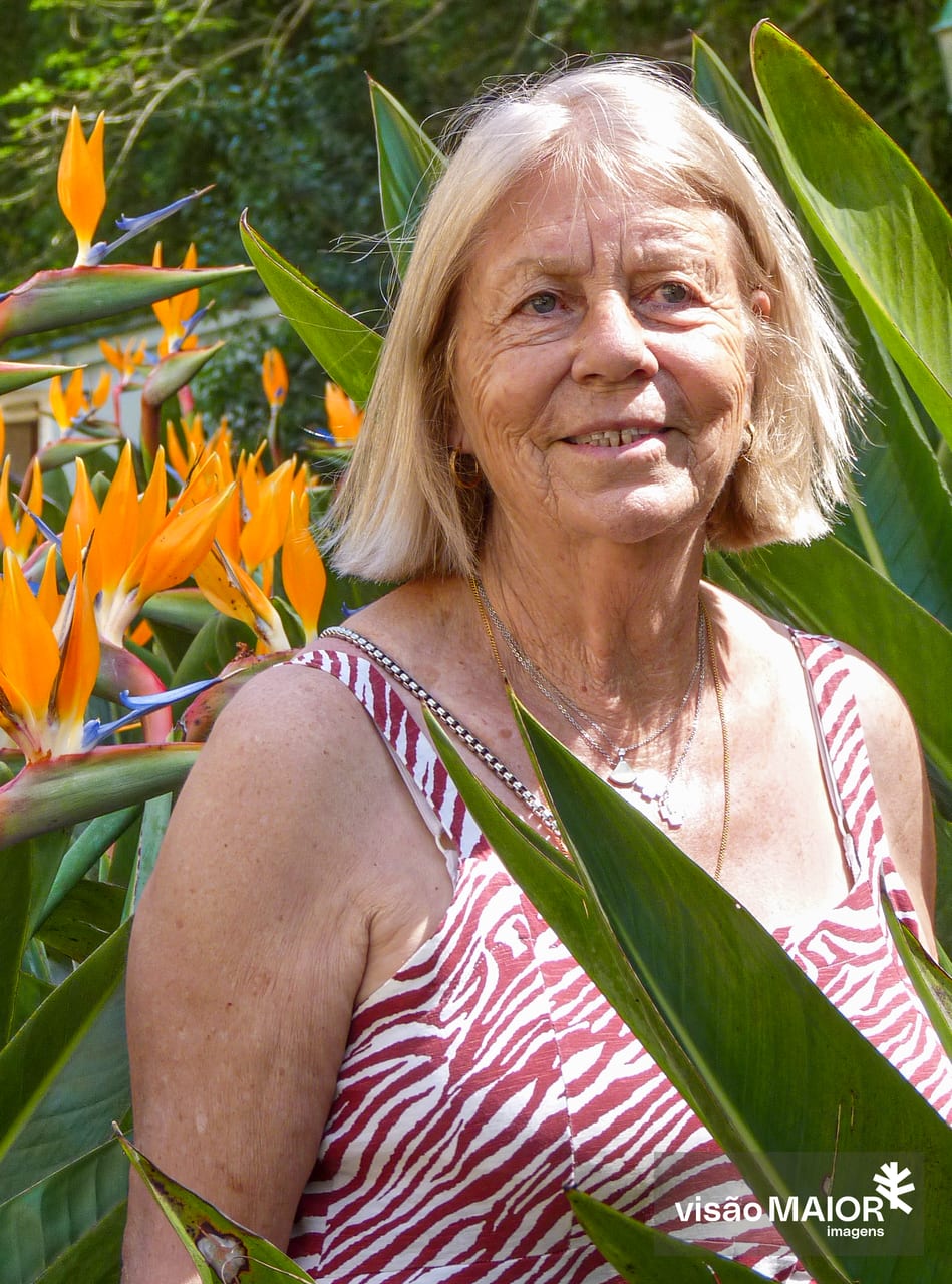 Mulher de cabelo e olhos claros, no meio de flores, com vestido de mangas à cava