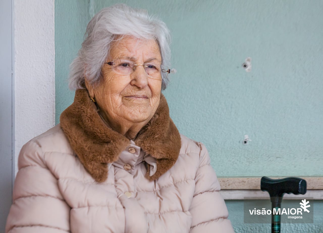 Mulher com cabelo claro e óculos, sentada à espera, com uma muleta e blusão claro com gola de pelo