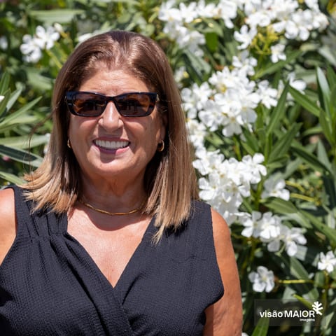 Mulher sorridente de cabelo castanho em pé, no meio de flores, de óculos escuros e top preto sem mangas