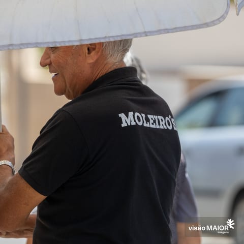 Homem de cabelo branco, de costas, a segurar um guarda sol, com t-shirt preta com um nome nas costas