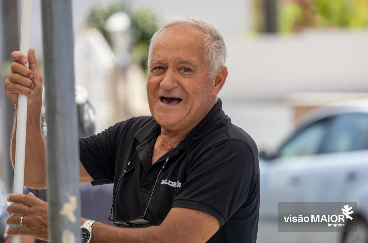 Homem de cabelos brancos e olhos claros, com os óculos caídos no peito a segurar um pau, sorridente