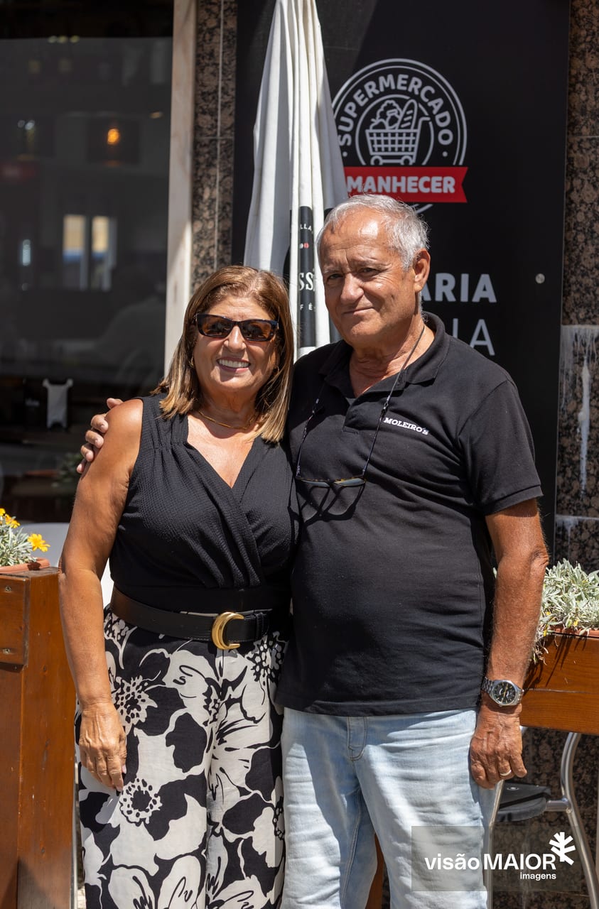 Casal abraçado em pé, ela de cabelos castanhos, ele de cabelos brancos, com um ar feliz e cumplice