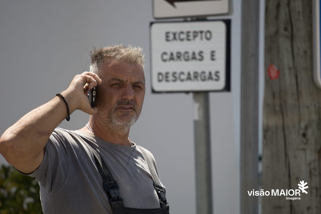 Homem de cabelo e barba grisalhos, macacão e t-shirt a falar telemovel com uma expressão apreensiva em frente a um sinal de trânsito