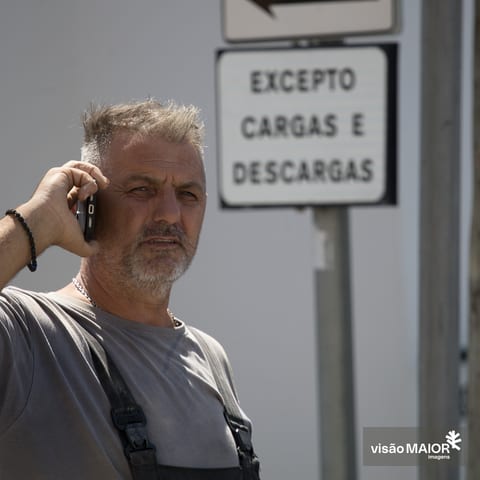 Homem de cabelo e barba grisalhos, macacão e t-shirt a falar telemovel com uma expressão apreensiva em frente a um sinal de trânsito