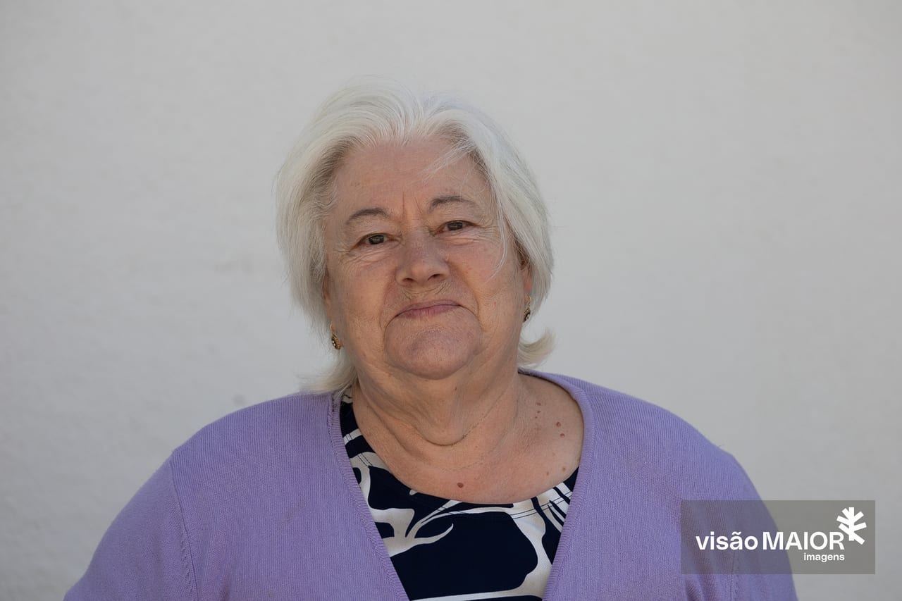 Mulher de cabelos brancos e olhos castanhos, brincos, casaco roxo e camisa às flores