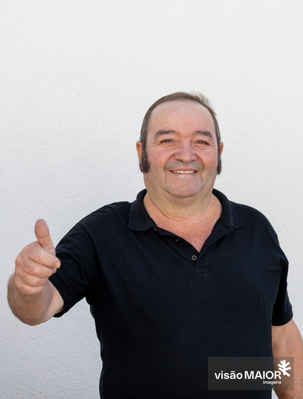 Homem de cabelo castanho com patilhas, em pé a rir a fazer um gesto com a mão