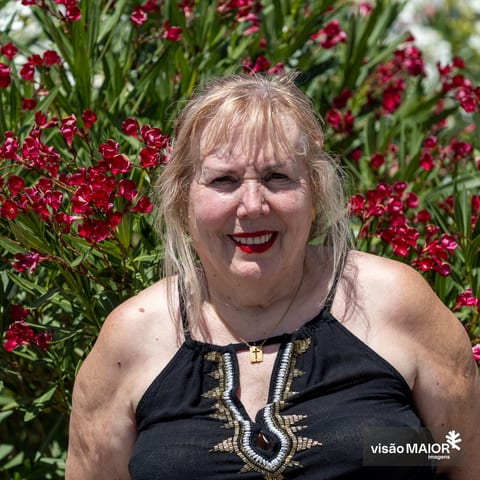 Mulher na rua, em frente a umas flores, cabelo claro e lábios pintados de vermelho, com top preto sem alças