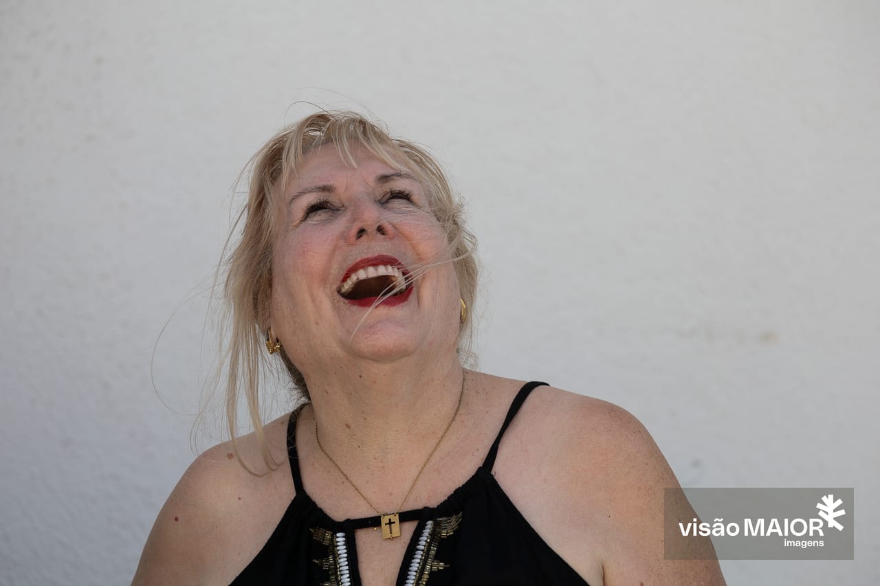 Mulher de cabelos claros e lábios ointados de vermelho, com um top preto, em fundo branco a olhar para cima a sorrir