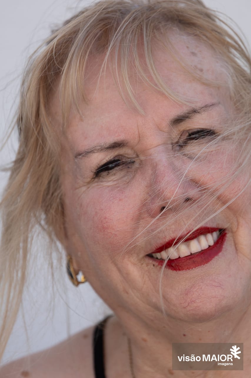 Mulher com cabelo claro a esvoaçar na cara, olhos castanhos e labios pintados, cabeça um pouco inclinada