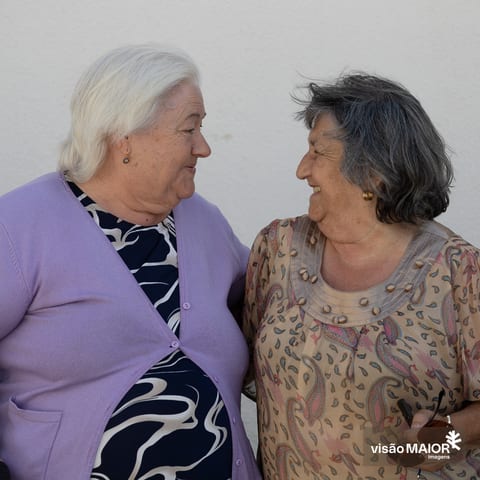 Duas mulheres de perfil, voltadas uma para a outra a rirem. Uma de cabelo preto, outra de cabelo branco