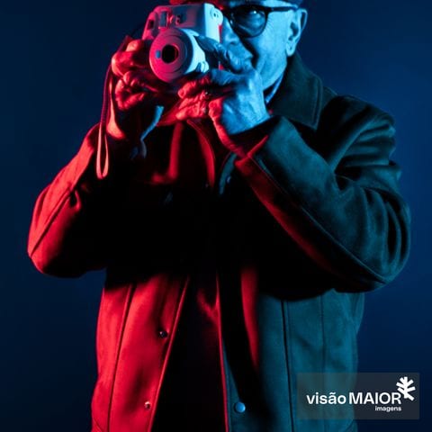 Homem de gorro, óculos e boné a tirar fotografias. Fundo escuro e reflexos vermelhos e azuis sobre ele.