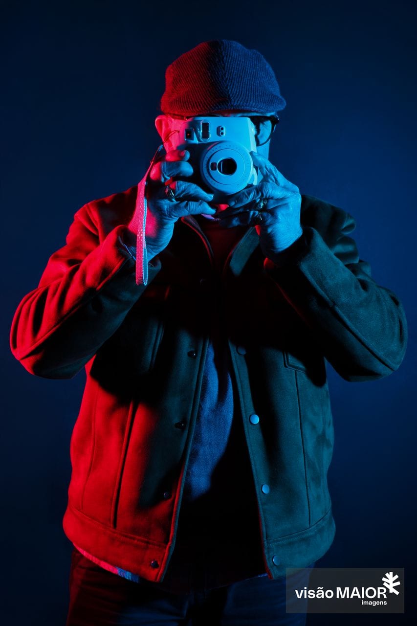 Homem de frente para câmara a tirar fotografias, com reflexos vermelhos e azuis, abeis nos dedos e blusão