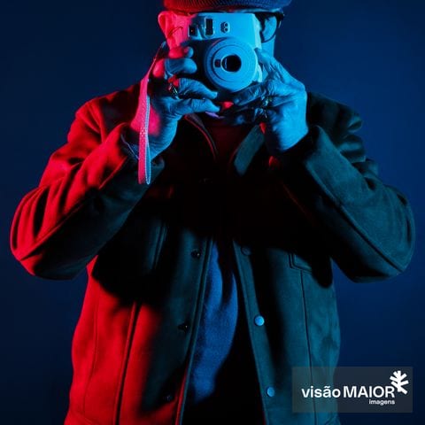 Homem de frente para câmara a tirar fotografias, com reflexos vermelhos e azuis, abeis nos dedos e blusão