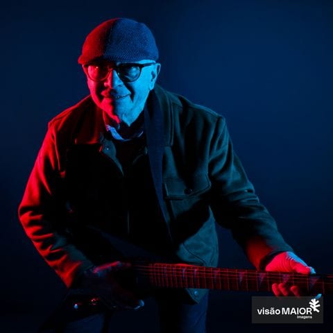 Homem de óculos, boné e blusão a tocar guitarra em fundo preto e com reflexos vermelhos e azuis