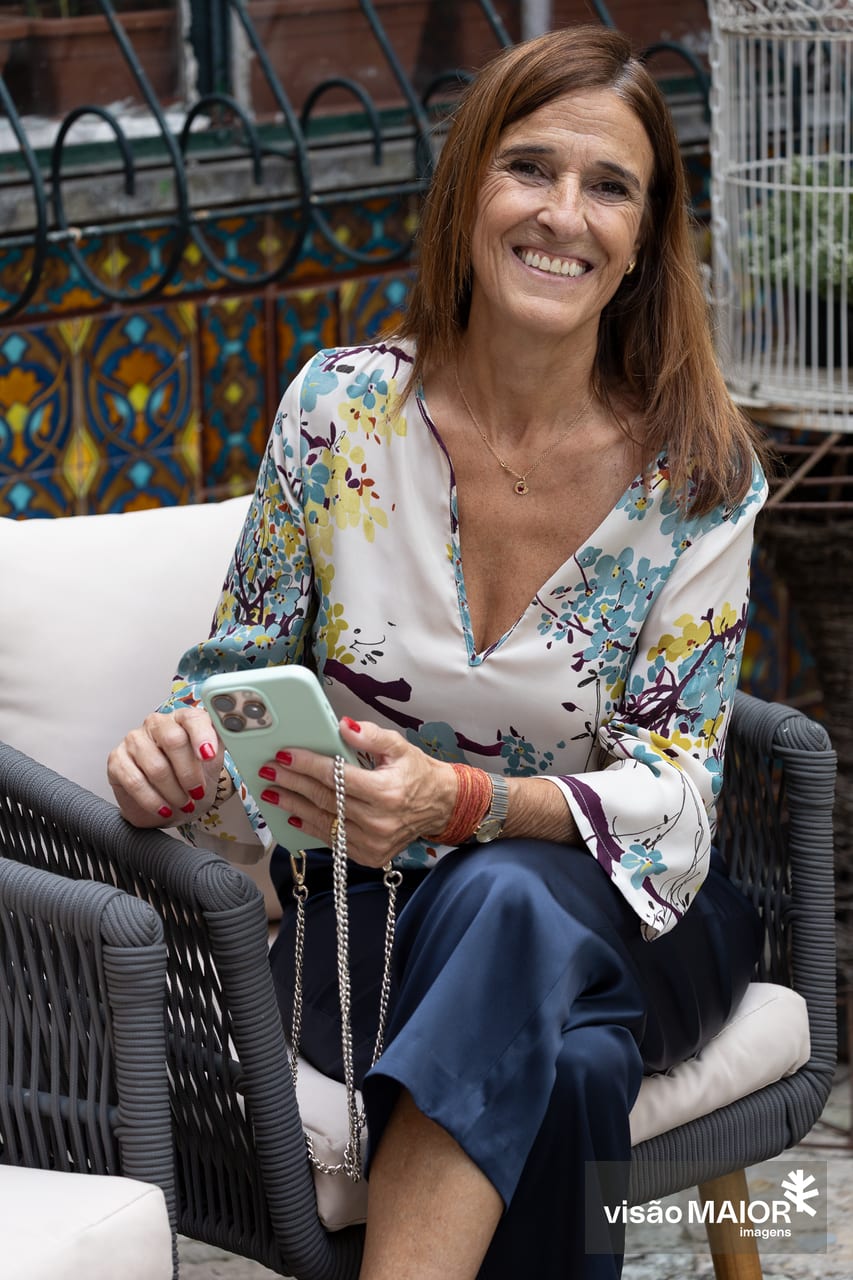 Mulher sentada, cabelos castanhos, camisa às flores e calças azuis, com um telemóvel na mão a sorrir