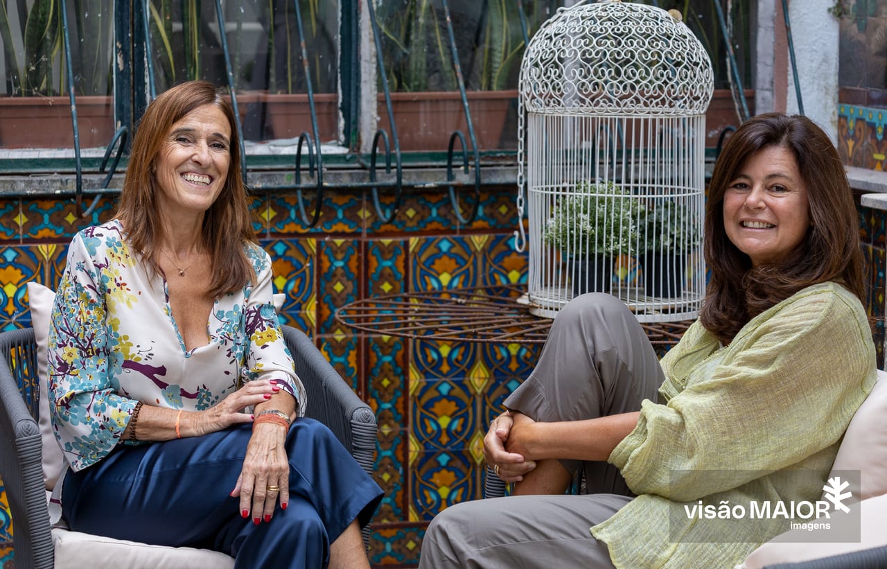 Duas mulheres de cabelo castanho, sentadas num páteo de azulejos a sorrirem, uma com uma perna levantada