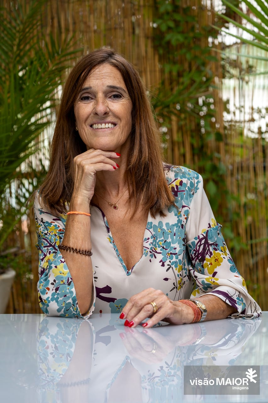 Mulher de olhos e cabelo castanho, camisa às flores, bricos, fio e anéis, com a mão no queixo a olhar para câmara