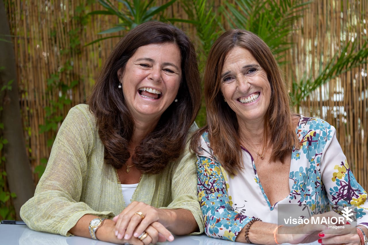 Duas mulheres ao ar livre, ambas de cabelo e olhos castanhos, com uma cerca de palhinhas e flores por trás a sorrirem