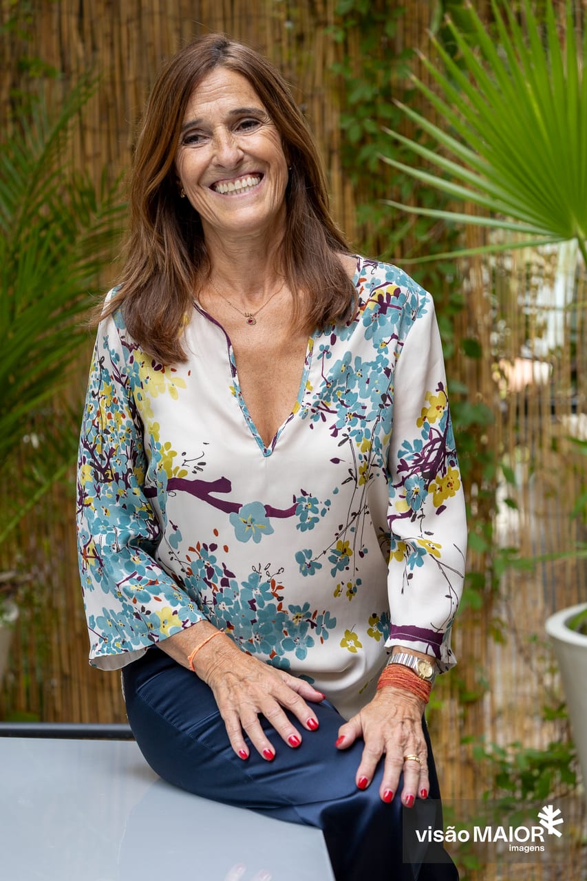 Mulher de cabelos e olhos castanhos, sentada de lado numa mesa, unhas pintadas, relógio, pulseira e fio a sorrir