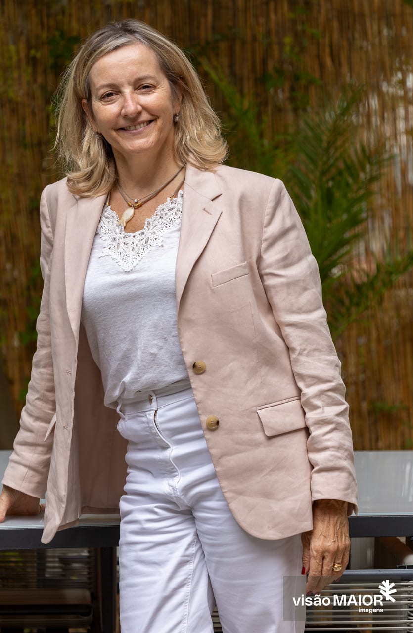 Mulher loira, vestida com roupa clara, em pé, com uma mão apoiada numa mesa, ao ar livre