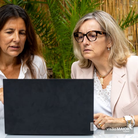 Uma mulher de cabelo loiro e outra de cabelo castanho e óculos, sentadas ao ar livre a olharem oara um computador